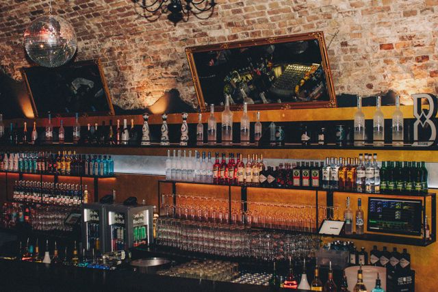 Die Bar im Eiskeller: beleuchtete Glasregale vor dem Backsteingewölbe, Discokugel und Goldrahmen-Spiegel