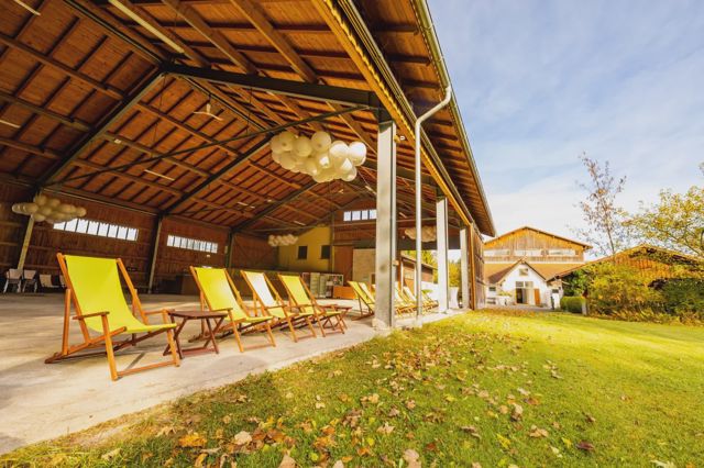 Die Maschinenhalle mit offenem Holzdachstuhl, zur Wiese hin offen, mit gelben Liegestühlen und Ballontrauben unter der Decke