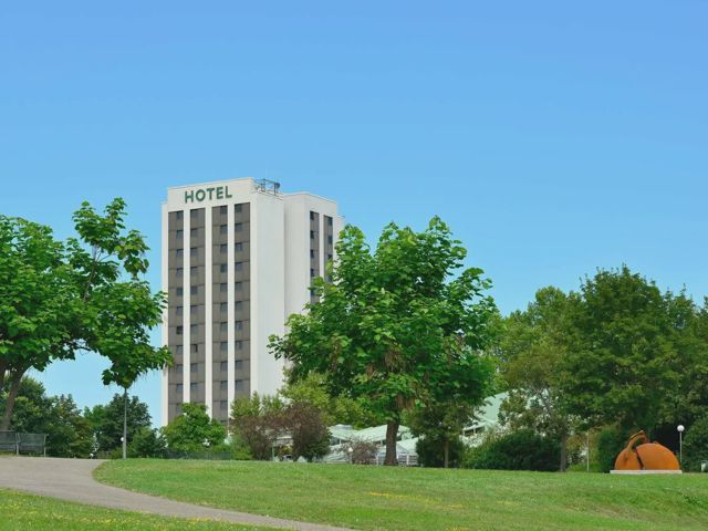 Das Hotel von außen: ein heller Turm mit dem Schriftzug HOTEL über Baumkronen, davor eine Wiese