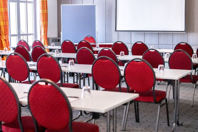 Parlamentarische Bestuhlung mit roten Stühlen, Pinnwand und Leinwand vor einer hellen Holzwand