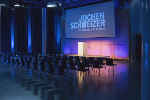 Bestuhlter Saal mit Bühne, Rednerpult und großer Leinwand, in blauem und orangem Bühnenlicht
