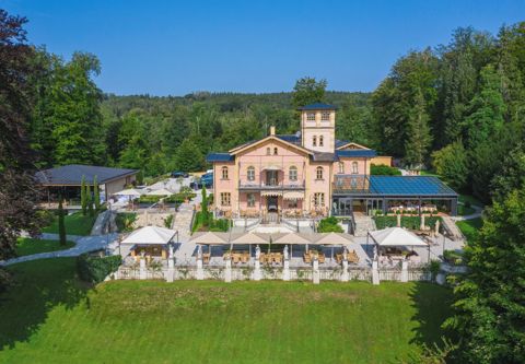 Die Villa von oben: rosafarbener Bau mit Turm, gläserne Orangerie, Terrassen mit Sonnenschirmen und Park bis zum See