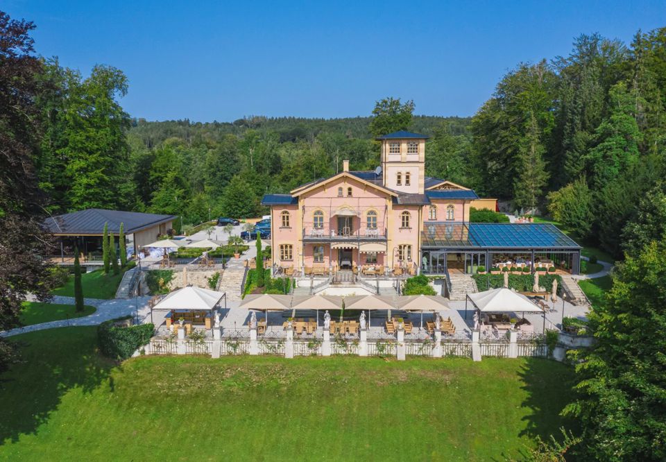 Die Villa von oben: rosafarbener Bau mit Turm, gläserne Orangerie, Terrassen mit Sonnenschirmen und Park bis zum See
