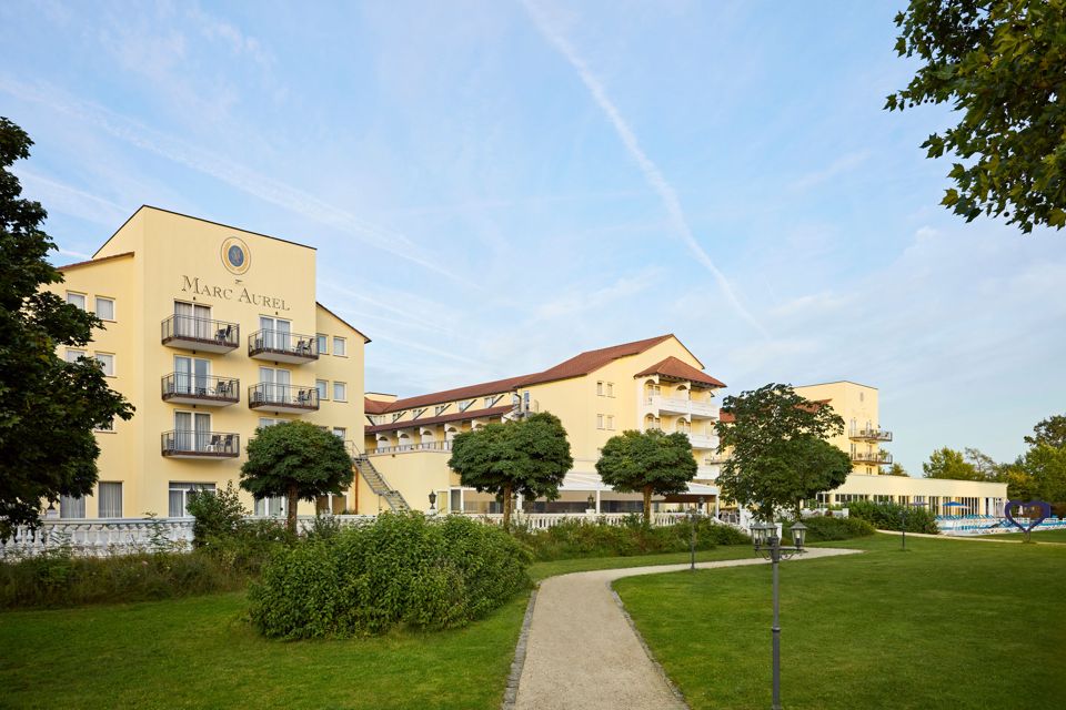 Das Haus vom Park aus: ein gelber Bau mit Balkonen und dem Schriftzug MARC AUREL, davor Rasen, Wege und geschnittene Bäume