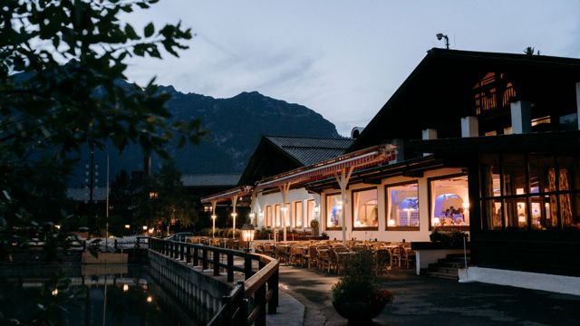 Das Hotel am Abend: beleuchtete Fensterfront mit gedeckten Tischen, davor die Terrasse am Wasser, dahinter die Bergkette