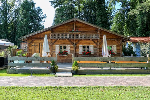 Die Alm am Chiemsee: eine Blockhütte mit geschnitztem Balkon, Geranienkästen und Fensterläden, davor eine Terrasse mit Biergartenbänken und Sonnenschirmen