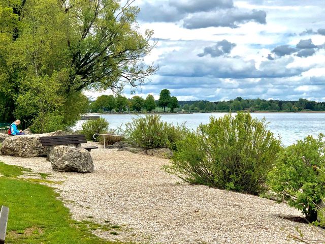 Das Kiesufer am Chiemsee mit Bank unter Bäumen, Blick über den See zum bewaldeten Ufer