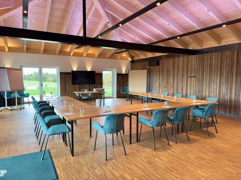 Der Bungalow unter offenem Holzdachstuhl: türkisfarbene Polsterstühle in U-Form an hellen Holztischen, dunkle Holzvertäfelung und Glastüren auf die Wiese